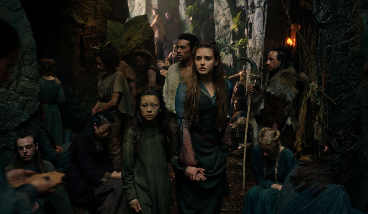 Nimue e Arthur attraversano una folla di persone in un villaggio alberato