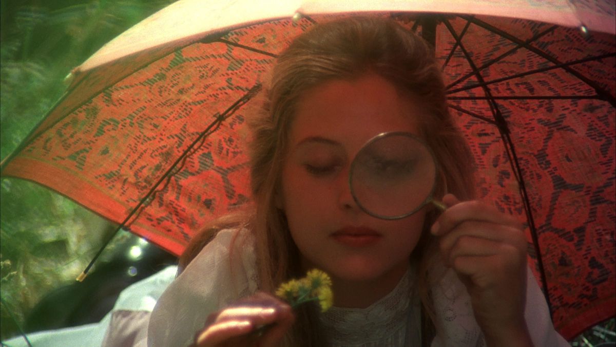 Una donna sotto un ombrello arancione brillante guarda attraverso una lente di ingrandimento in Picnic a Hanging Rock