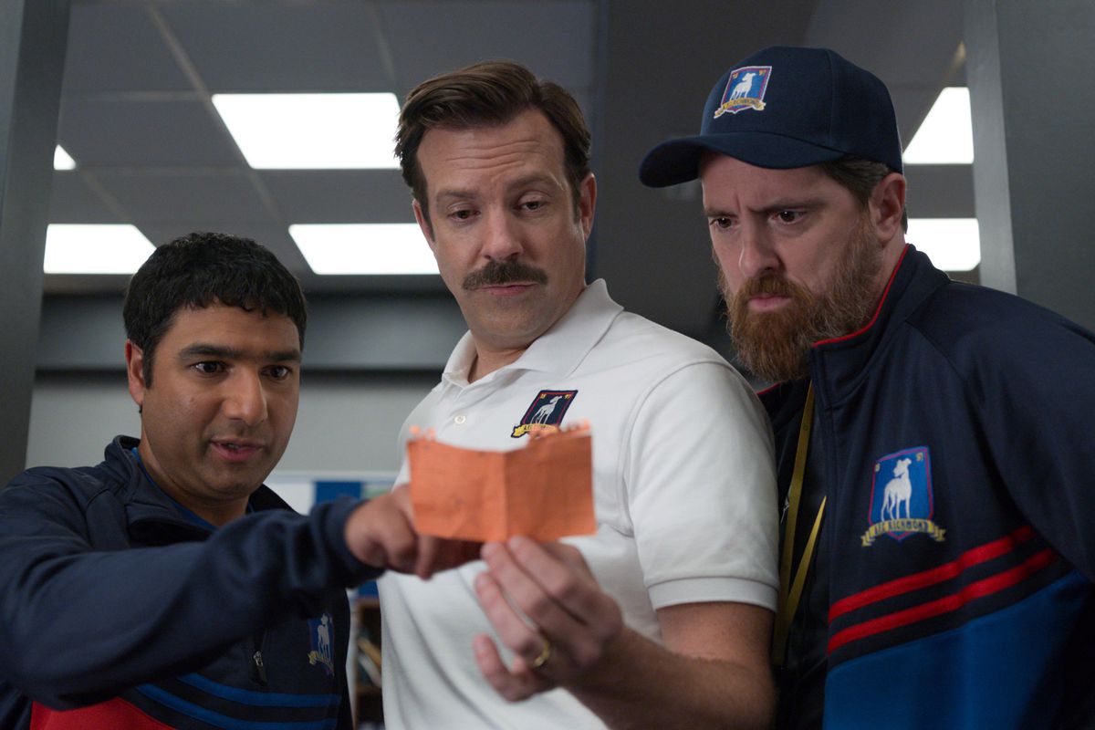 Ted Lasso and his assistant coaches lean over a note