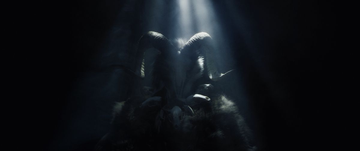 una minacciosa figura ammantata che indossa un teschio di ariete come maschera si trova sotto un inquietante raggio di luce.