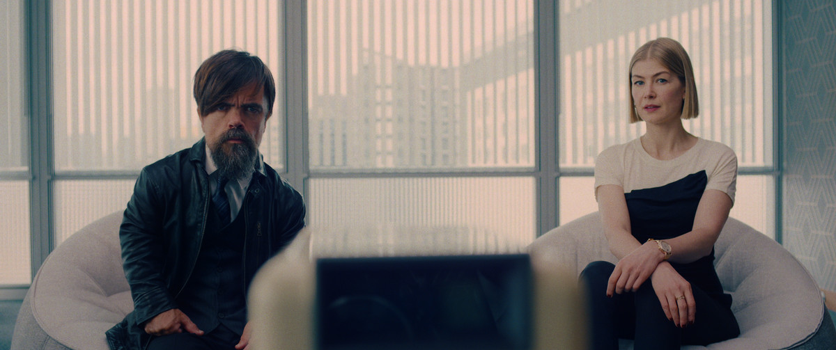 Peter Dinklage e Rosamund Pike siedono insieme su sedie davanti a una finestra luminosa, guardando qualcosa su uno schermo in primo piano in I Care a Lot