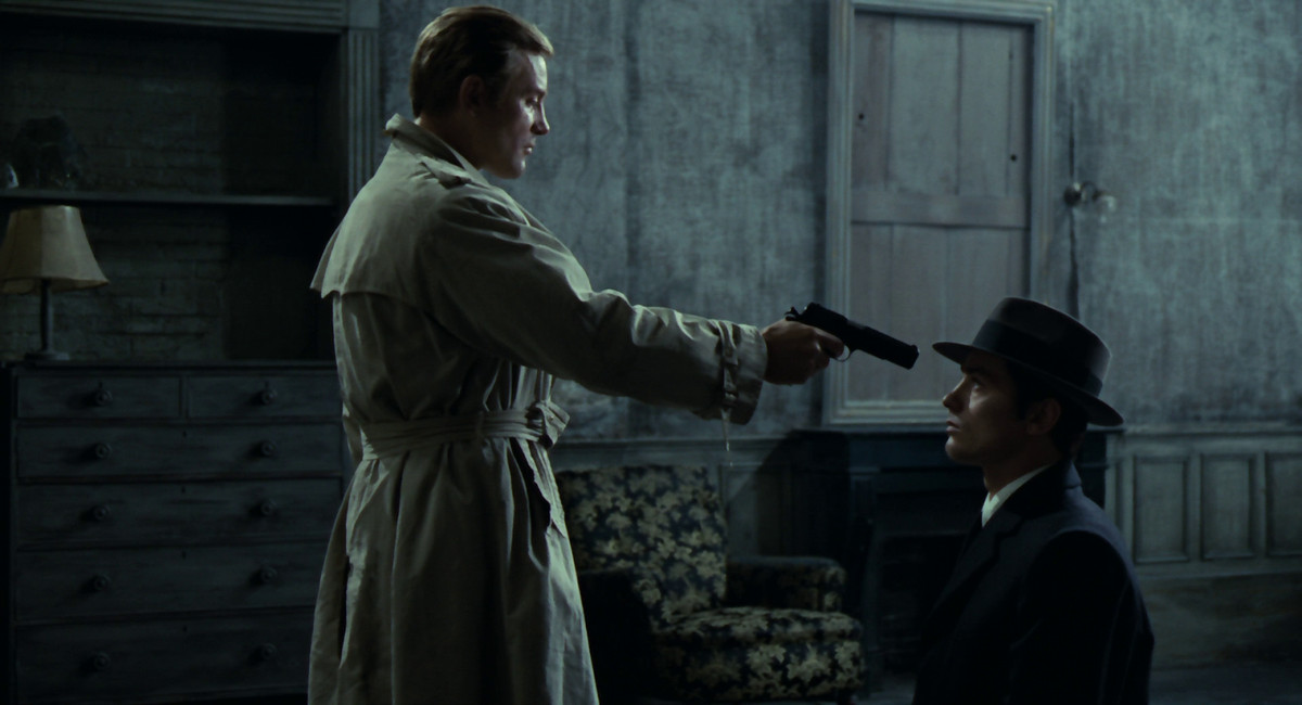 Alain Delon nei panni di Jef Costello siede immobile mentre un detective gli punta una pistola in faccia.