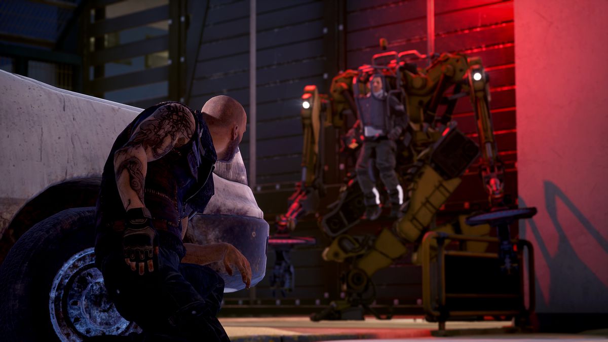 Un uomo calvo, muscoloso e tatuato si accovaccia accanto a un'auto, osservando un uomo più piccolo all'interno di un grande scheletro meccanico