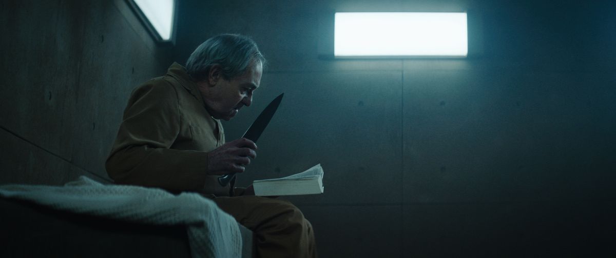 An elderly man clutching a nasty-looking knife and reading a book sits on his bed in a dimly lit concrete cell in The Platform.