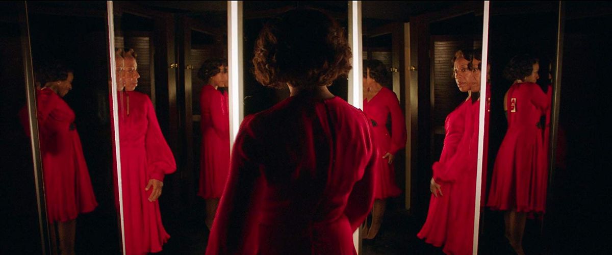 Una donna in un abito rosso vivo si trova in mezzo a una serie di specchi che riflettono se stessa e l'un l'altro in In Fabric