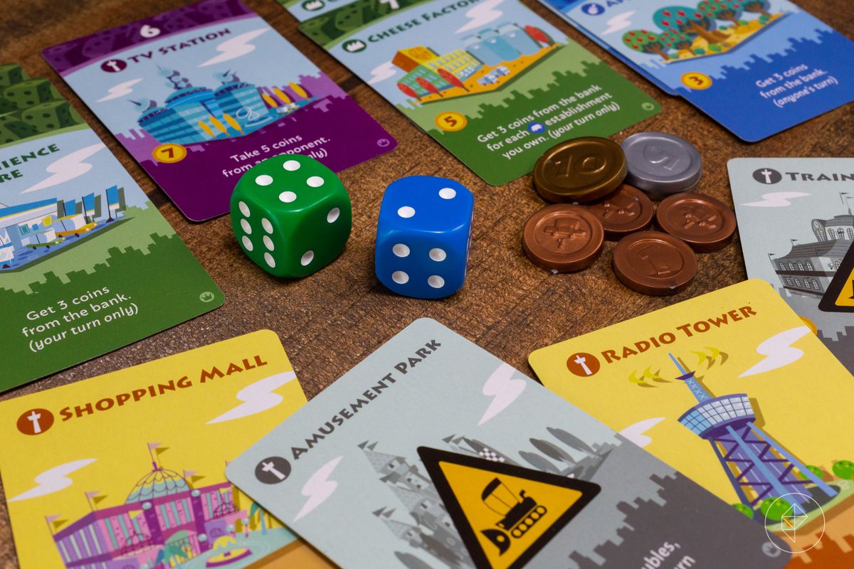 A pile of bits from Machi Koro, including a shopping mall, an amusement park, and a radio tower. Two dice and some coins sit in the center of the frame.