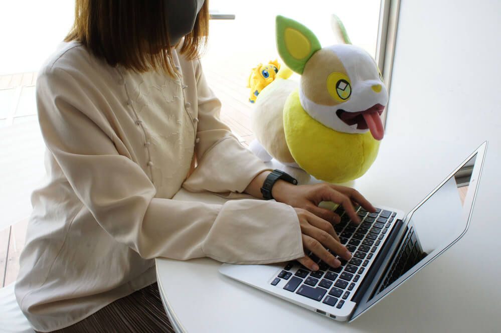 Una donna lavora su un laptop accanto a un peluche di Yamper con Joltik attaccato al sedere