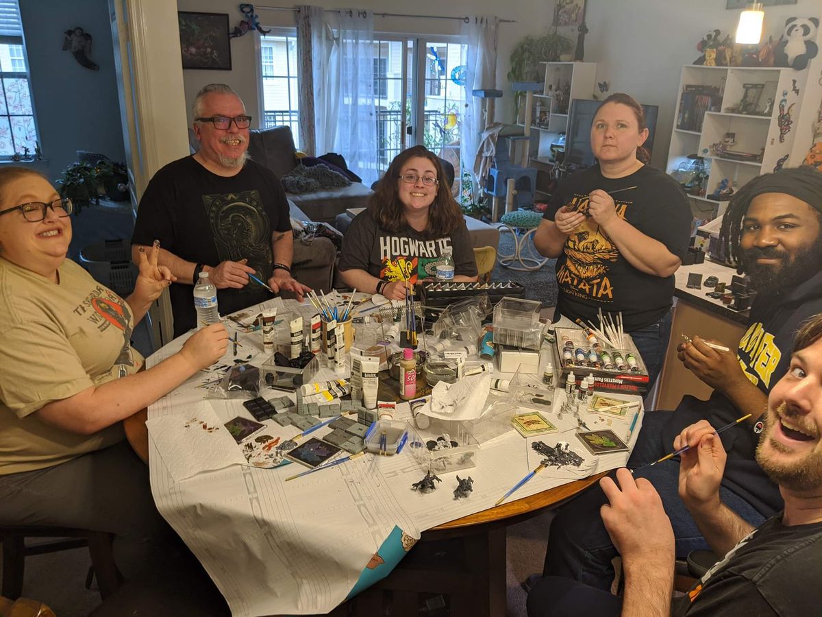 Un gruppo di giocatori siede intorno a un tavolo ricoperto di carta da pacchi. I colori sono sparsi e le miniature sono in lavorazione.