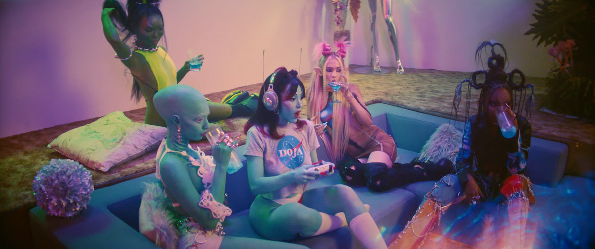 Doja Cat siede in un futuristico soggiorno incassato in stile metà del secolo mentre tiene in mano un controller per PlayStation 5. è circondata da un sacco di ragazze aliene