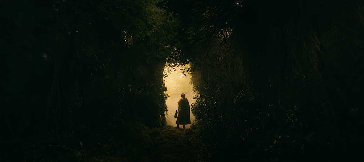 Gawain (Dev Patel) si staglia contro una luce ambrata mentre guarda attraverso la foresta oscura davanti a sé in The Green Knight
