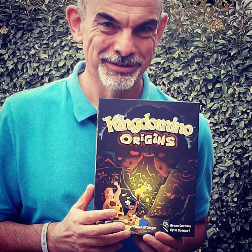 Davanti a una siepe c'è un uomo con il pizzetto sale e pepe.  Nelle sue mani una copia di Kingdomino Origins, con un grande vulcano sul davanti.