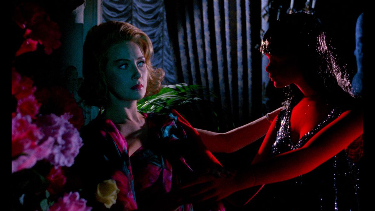 Una donna in una stanza scarsamente illuminata, circondata da vividi fiori rosa, guarda con diffidenza un manichino rosso brillante con lunghi capelli neri e un vestito nero