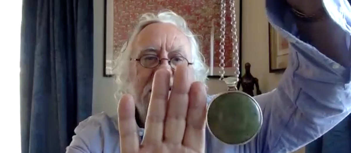 Dan Hennah holds up a round pendant on the end of a bulky chain, next to his hand to show scale. The green stone set in it is larger than the first two knuckles of his pointer finger.