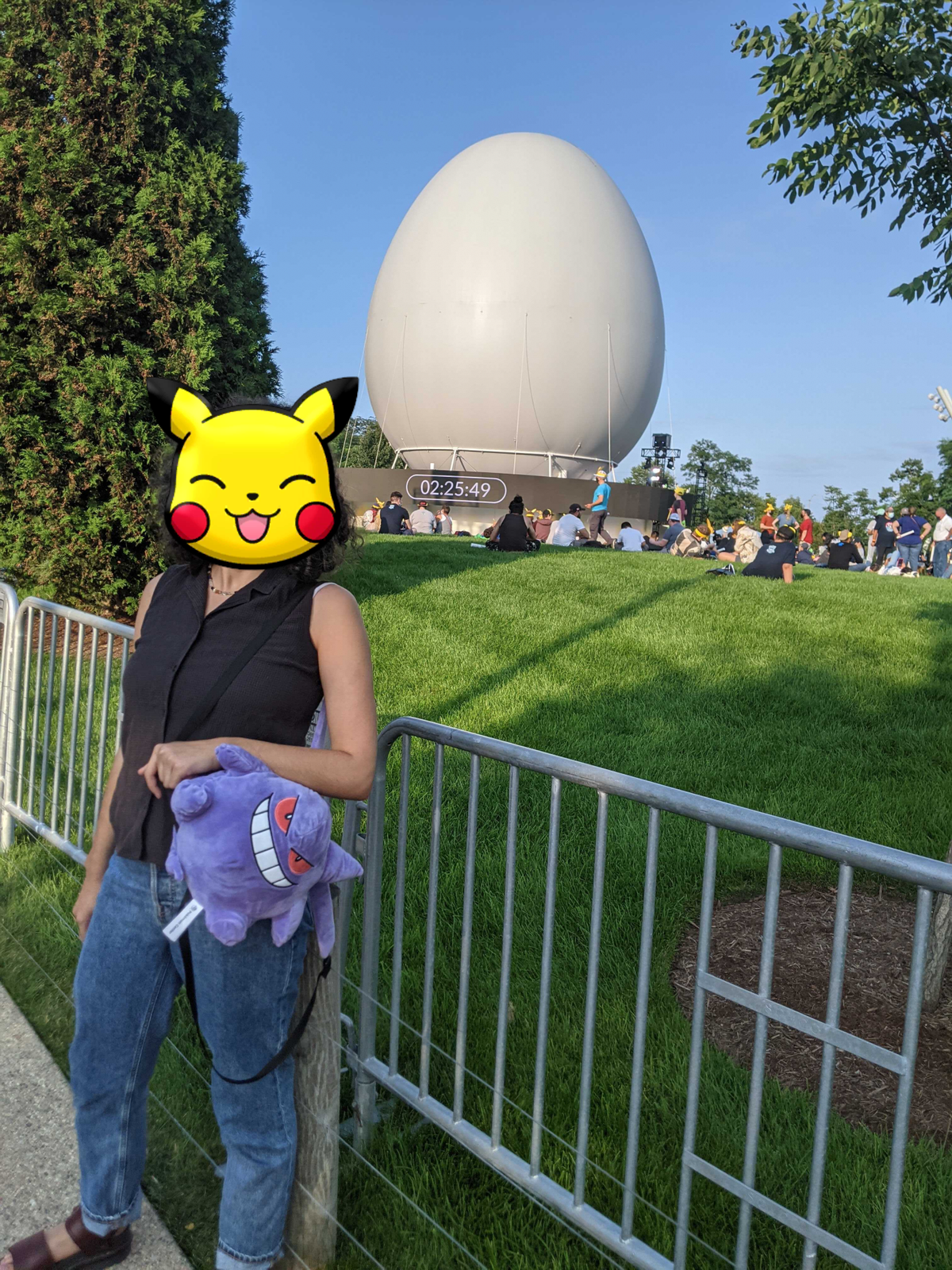 una persona in piedi al pokemon go fest che indossa uno zaino gengar. la sua faccia è bloccata da pikachu.