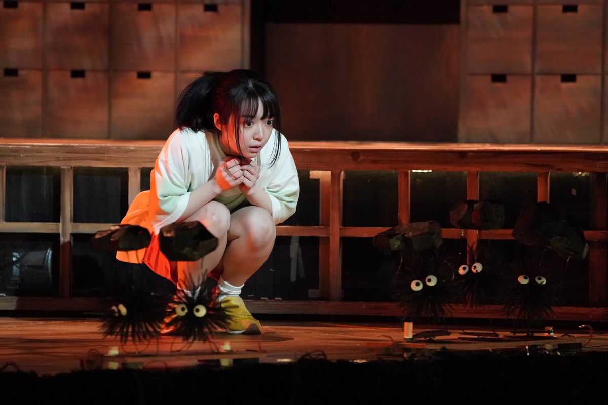 Chihiro si accovaccia e guarda le marionette di folletti di fuliggine che trasportano carbone che si muovono sul palco in Spirited Away: Live on Stage