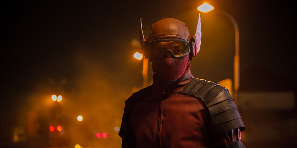 un uomo con una maschera rossa con le ali si trova su una strada nebbiosa illuminata da lampioni arancioni