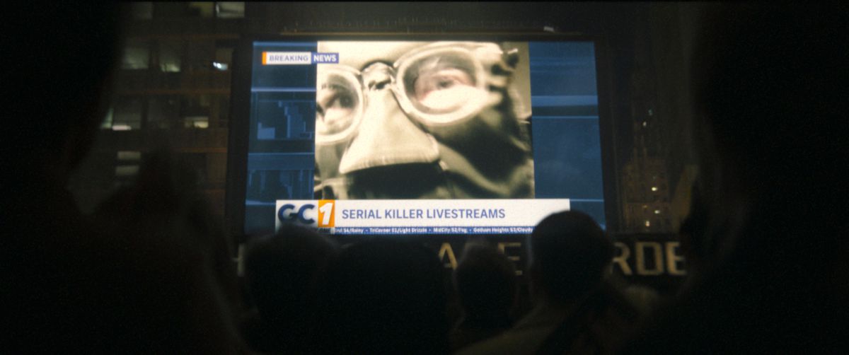 Gli spettatori di The Batman guardano il telegiornale, che mostra un primo piano della maschera dell'Enigmista e dei suoi spessi occhiali. Un chirone recita