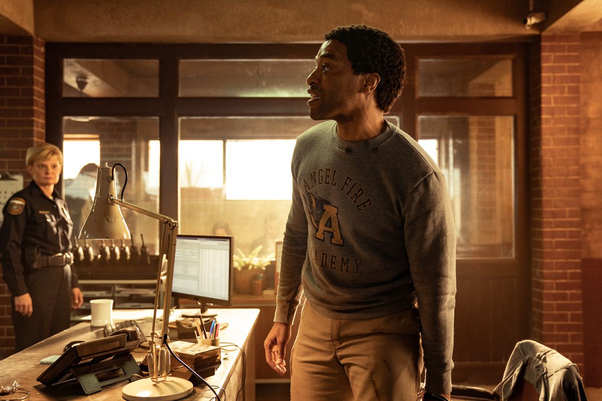 Faraday in piedi con indosso una felpa mentre un poliziotto guarda sullo sfondo