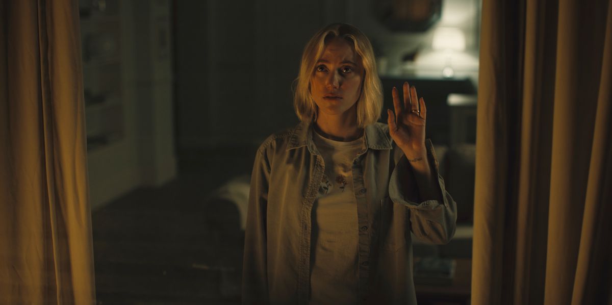 Maika Monroe è in piedi vicino a una finestra, guardando verso l'esterno, con la mano sul vetro in Watcher