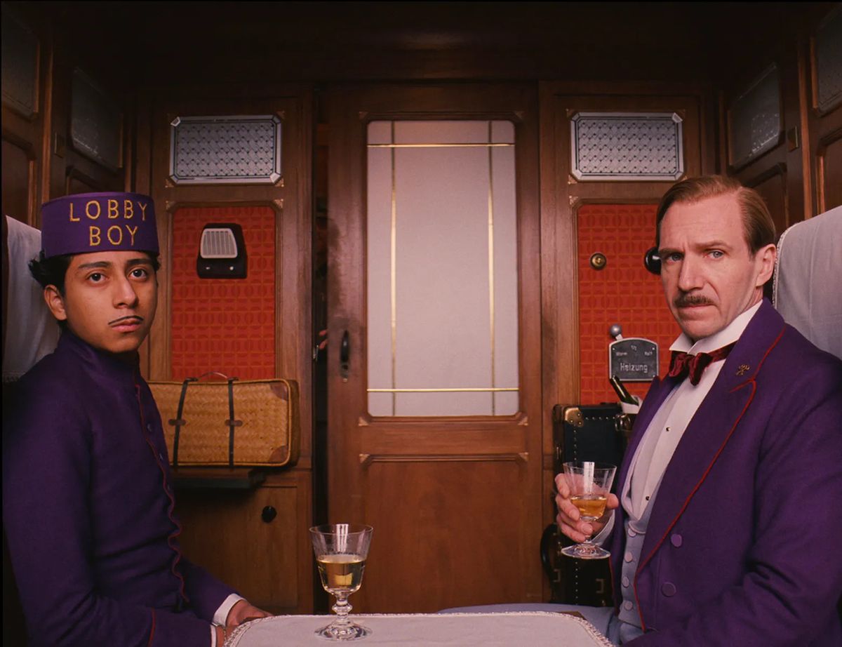 Ralph Fiennes e Tony Revolori indossano abiti viola abbinati in un vagone del treno al Grand Budaapest Hotel.