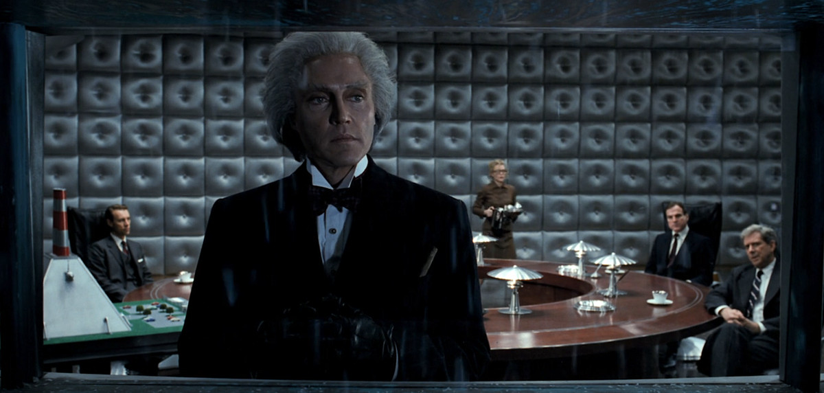 Max Schreck (Christopher Walken) è inquadrato in una finestra, a guardare una Gotham natalizia innevata con una sala riunioni piena di uomini compiaciuti alle sue spalle, in Batman Returns