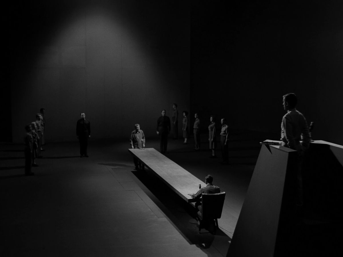 Un uomo è in piedi all'estremità di un tavolo in attesa di processo nell'episodio di The Twilight Zone