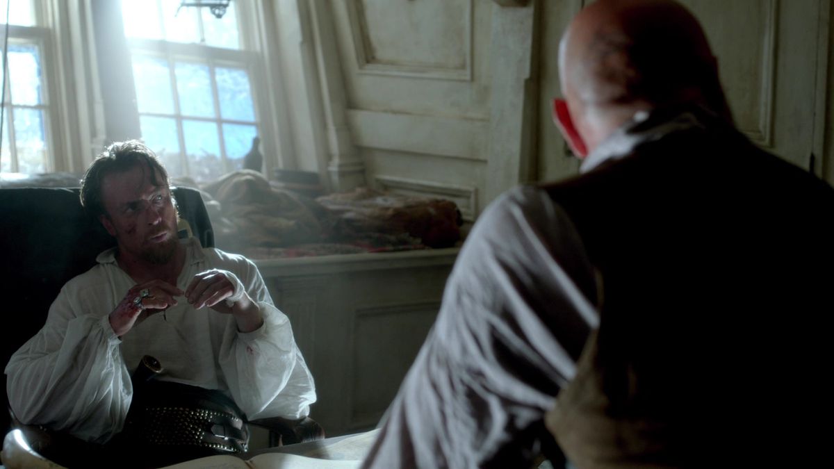 Il capitano Flint seduto alla sua scrivania mentre un altro pirata gli parla