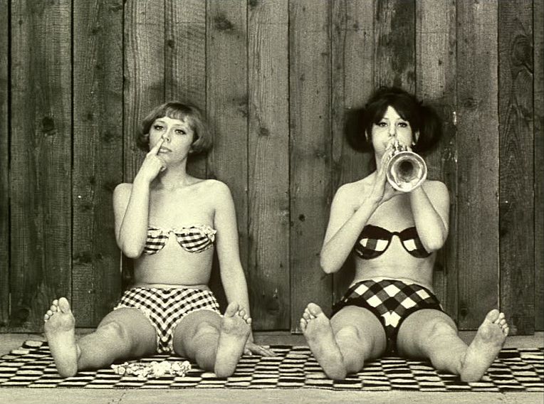 Le due star di Daisies oziano in costume da bagno, una con il dito nel naso e l'altra che suona uno strumento d'ottone.