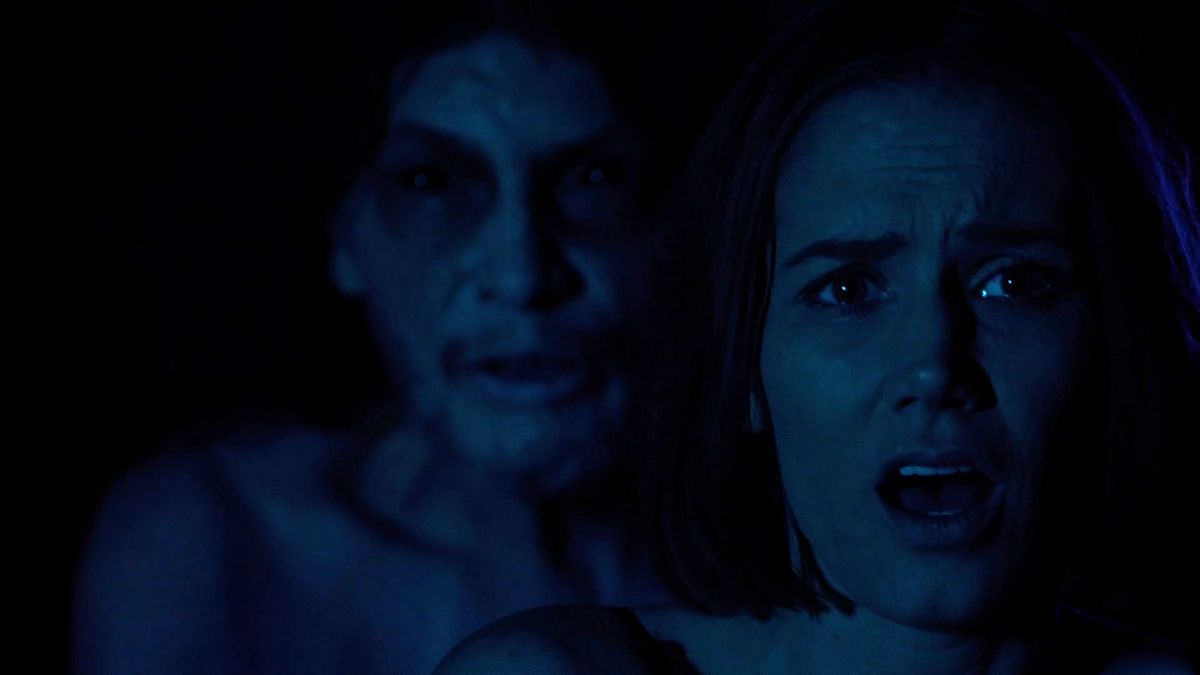 Una donna fissa con la bocca spalancata mentre una figura pallida e dall'aspetto spettrale si china sopra la sua spalla.