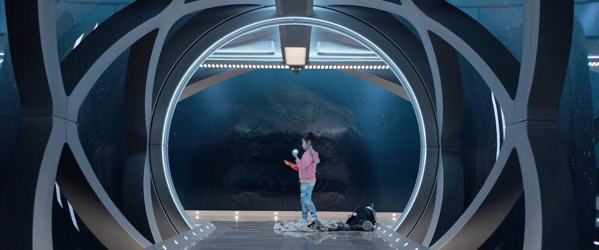 Uno squalo megalodonte fissa una ragazza che tiene una palla blu attraverso un tunnel di vetro in The Meg.