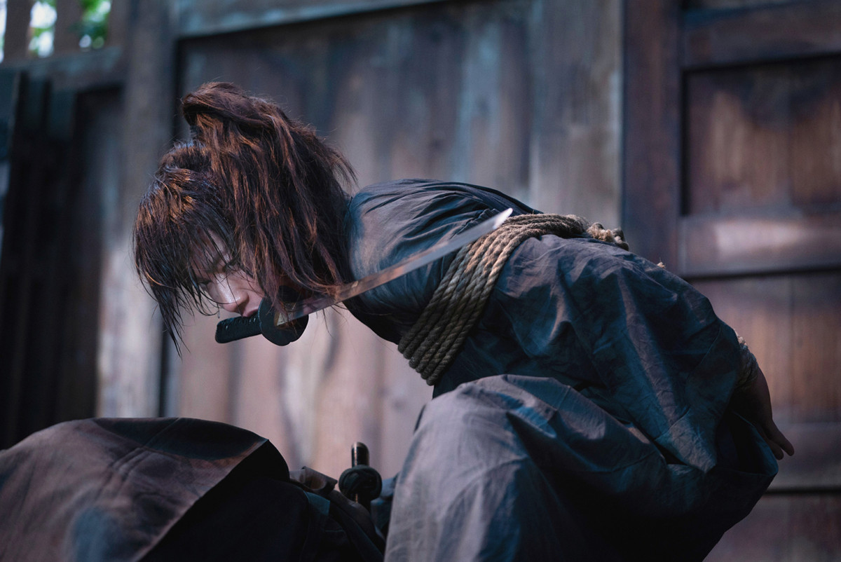 Takeru Satoh nei panni di Kenshin Himura che tiene una spada in bocca con le mani legate dietro la schiena in Rurouni Kenshin: The Beginning.