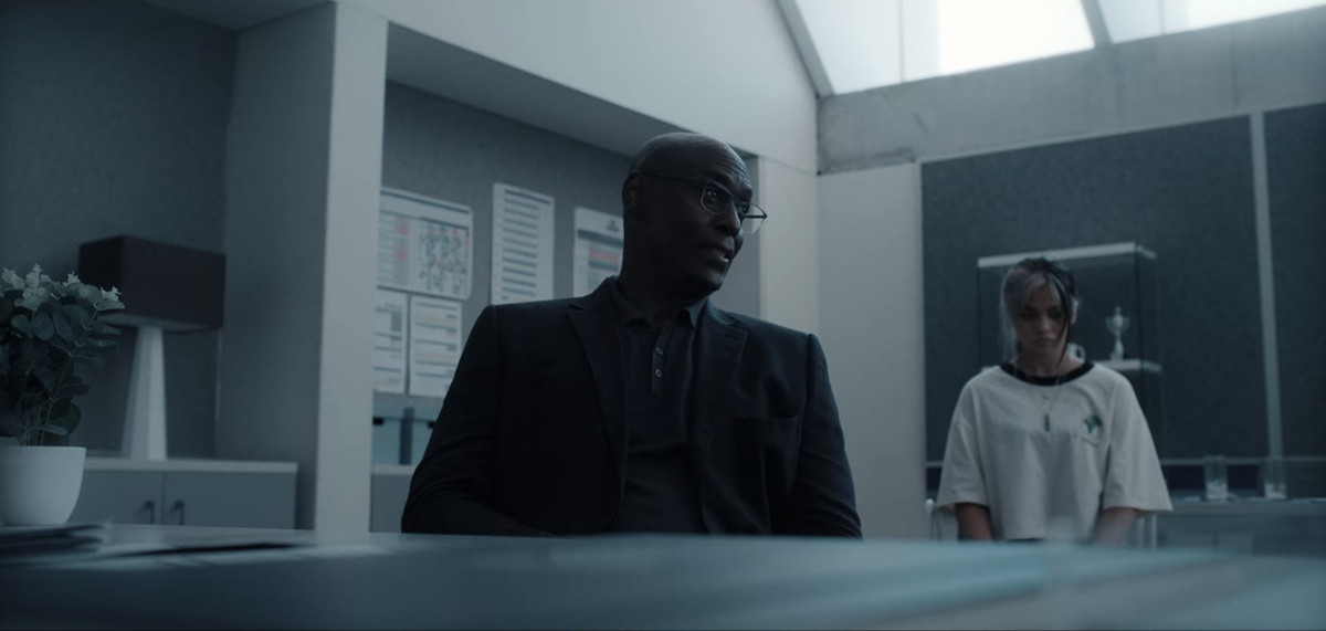 Albert Wesker seduto su una sedia con aria delusa mentre sua figlia siede dietro di lui con la testa leggermente china