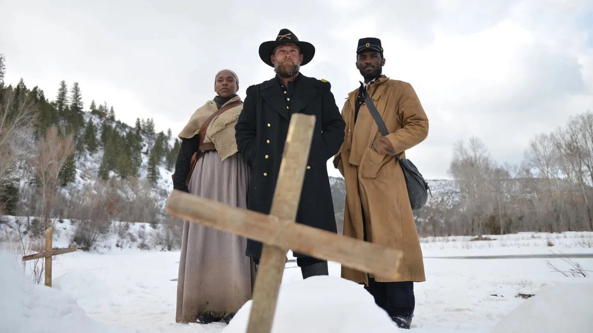 Un trio di pistoleri in trench sta davanti a una croce di legno circondata da un campo innevato.