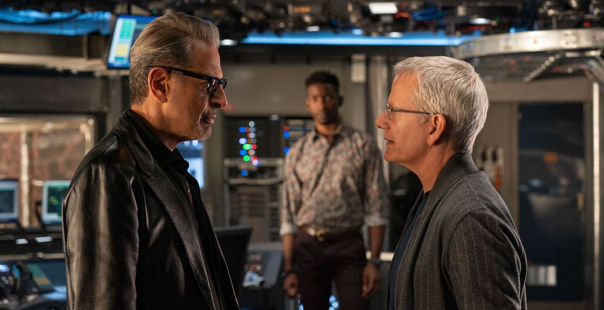 Il dottor Ian Malcolm (Jeff Goldblum) e Lewis Dodgson (Campbell Scott) si affrontano in primo piano in una stanza molto tecnologica, in stile pistolero, mentre Ramsay Cole (Mamoudou Athie) guarda sullo sfondo in Jurassic World Dominion.