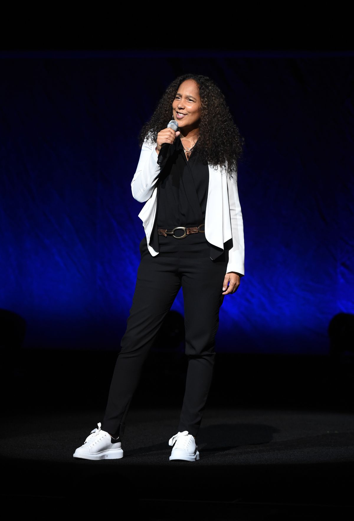 Gina Prince-Bythewood è in piedi sul palco davanti a uno skrim nero e blu con in mano un microfono e comandando il pubblico