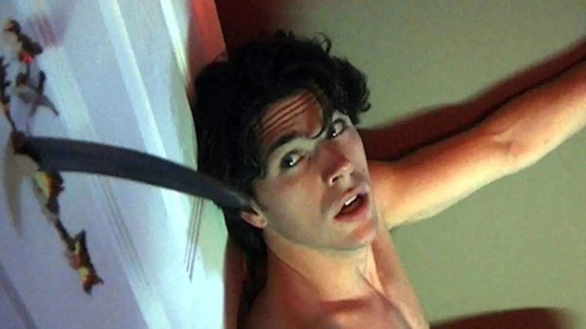 Un giovane uomo a torso nudo osserva con paura qualcuno che pugnala attraverso una porta a Hellbent.