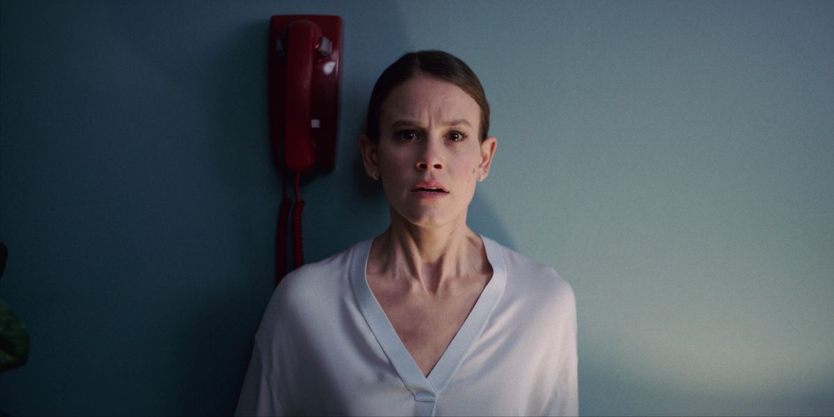 Rose (Sosie Bacon) indossa una camicetta azzurra e i suoi capelli castani tirati indietro sussulta mentre si appoggia al muro, in piedi accanto a un telefono rosso in Smile