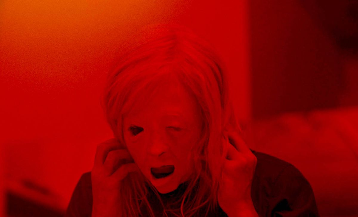 Una figura che indossa una maschera macabra di un volto di donna in Possessore, davanti a uno sfondo rosso brillante.