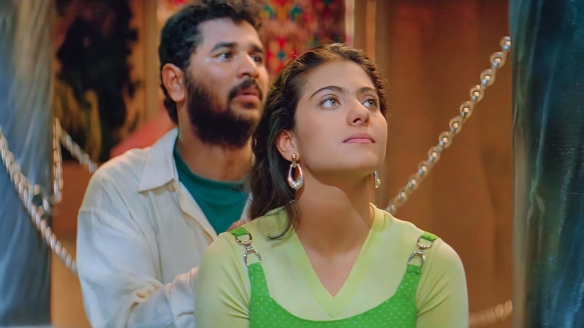 Una donna che indossa un top verde e un orecchino a cerchio fissa fuori da una finestra, mentre un uomo barbuto in piedi dietro di lei guardando nella stessa direzione a Minsara Kanavu.