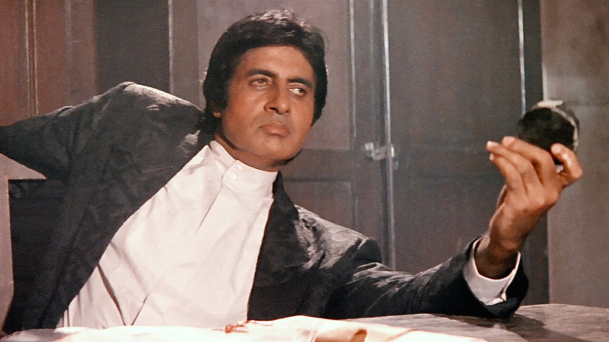 Amitabh Bachchan tiene in mano un disco nero rotondo mentre si appoggia su un tavolo e una sedia ad Agneepath.