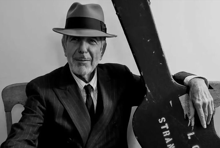Leonard Cohen, con indosso giacca e cravatta e un bel cappello, tiene una custodia per chitarra.