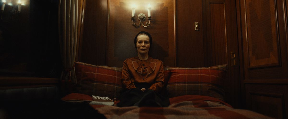 Una donna anziana si siede su un letto con un libro accanto a lei. Un paio di candele si accendono in alto.