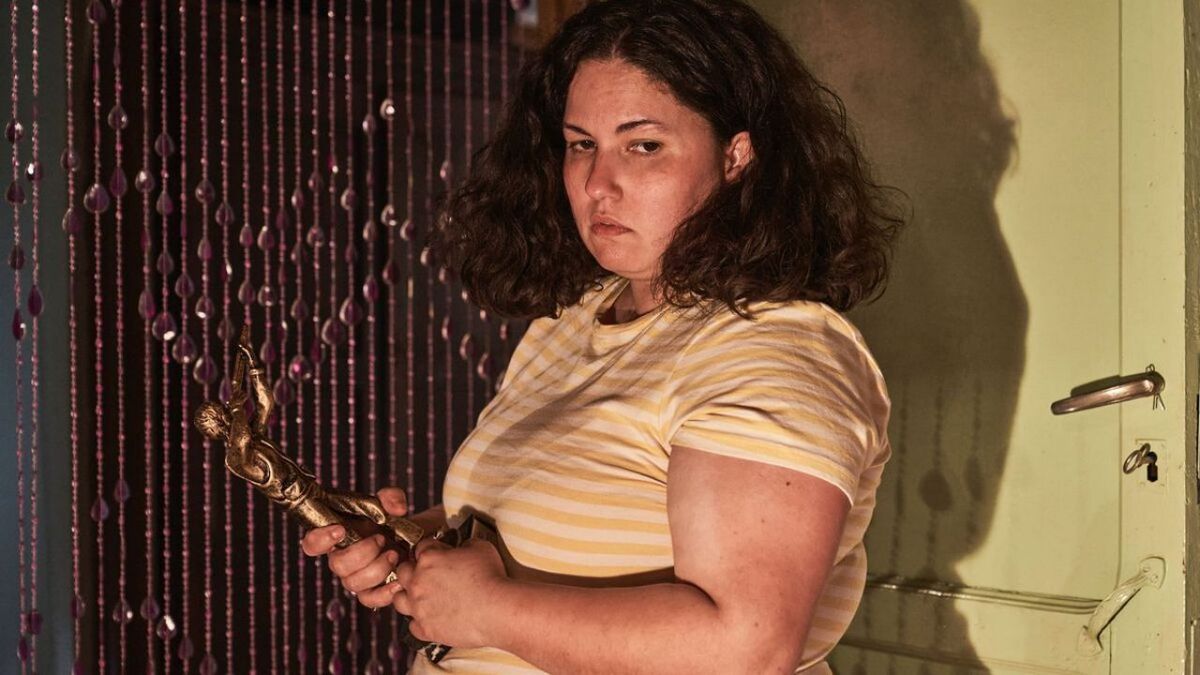 Una giovane donna indossa una camicia a righe e tiene una statua mentre è in piedi davanti a una tenda di perline e una porta a Piggy.