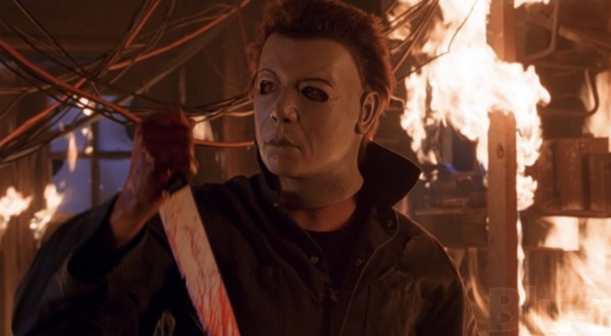 Michael Myers, circondato dalle fiamme, indossa una maschera con sopracciglia arcuate e un naso piรน distinto. Tiene in mano un coltello insanguinato.