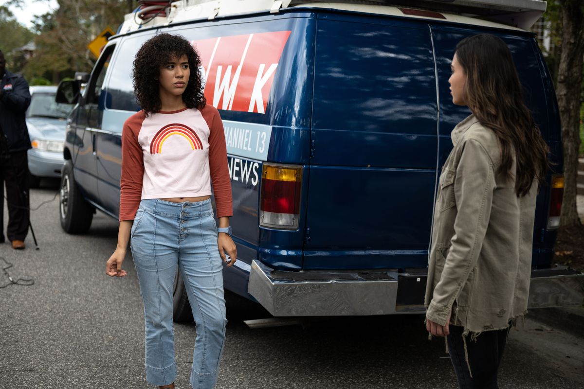 Jasmin Savoy Brown indossa una maglietta a maniche lunghe con un arcobaleno sopra e jeans davanti a un camion di notizie in Scream del 2022.