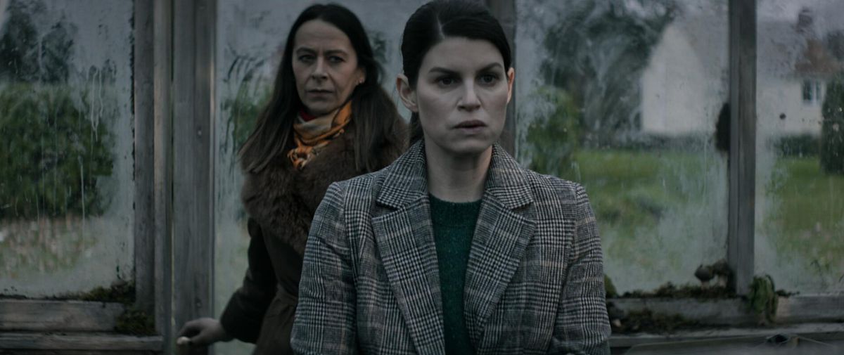 Una donna con una giacca di tweed grigio che indossa un maglione verde si trova all'interno di una serra con finestre opache di fronte a una donna anziana con una pelliccia marrone con una sottile sciarpa arancione.