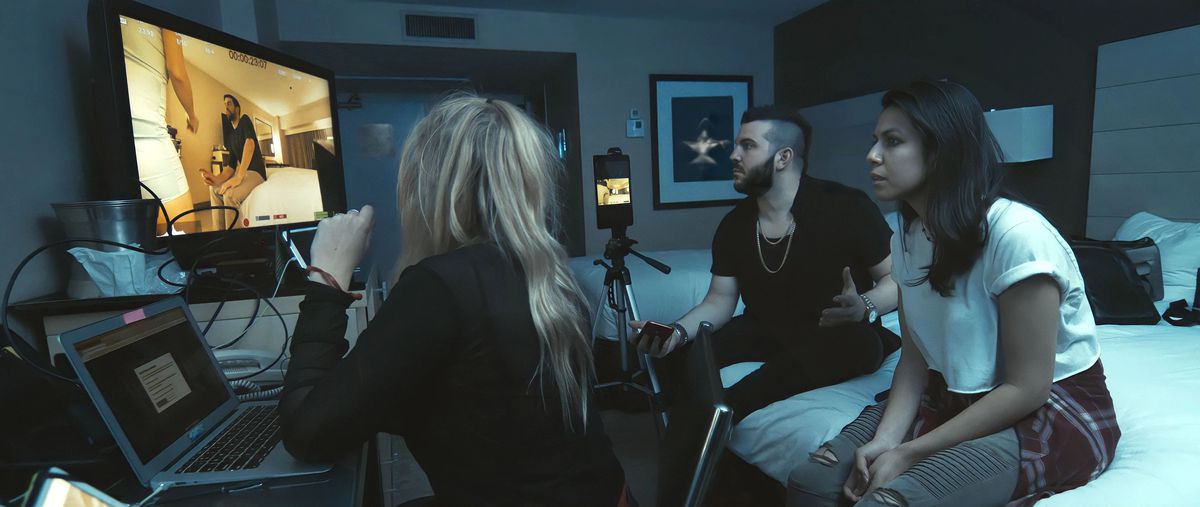Tre persone, un uomo e due donne, siedono in una stanza d'albergo buia davanti a uno schermo che mostra le riprese di una videocamera portatile.