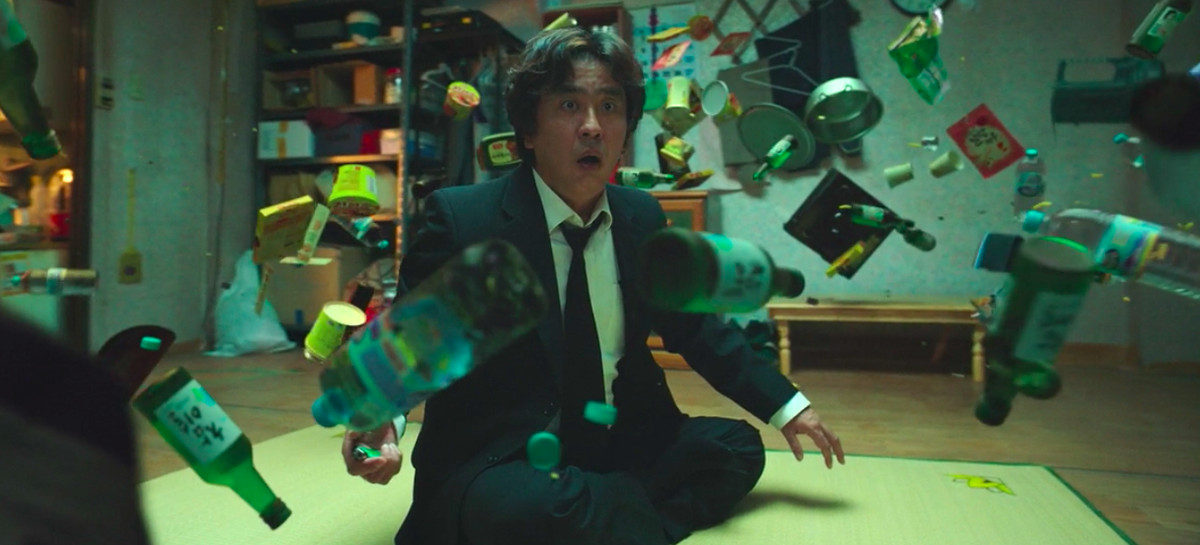 Oggetti domestici casuali galleggiano davanti a un uomo dall'aspetto sbalordito, che è seduto a gambe incrociate sul pavimento.