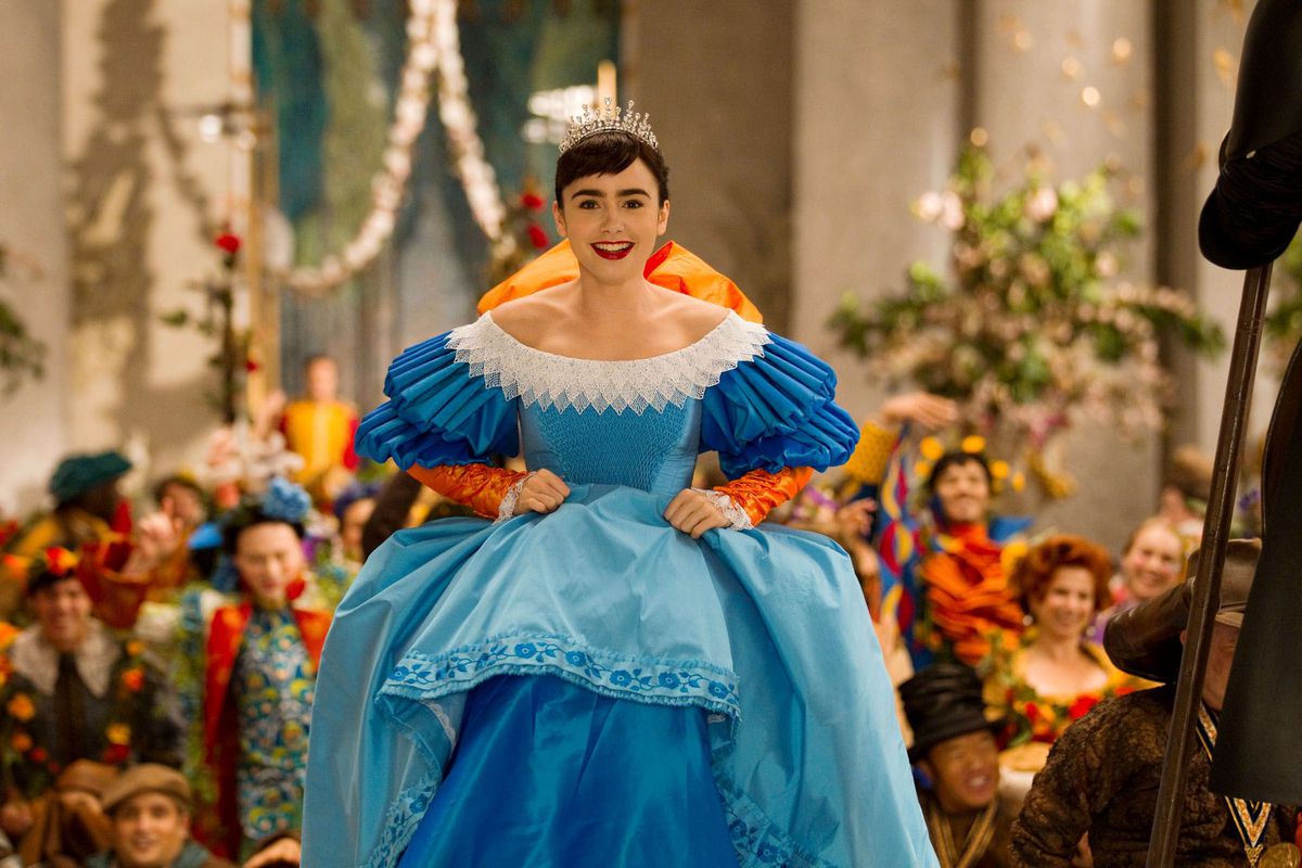 Biancaneve (Lily Collins) sale le scale della sala del trono con un sontuoso abito blu con rifiniture bianche e un gigantesco fiocco arancione sulla schiena mentre le persone vestite con costumi altrettanto colorati fanno il tifo per lei