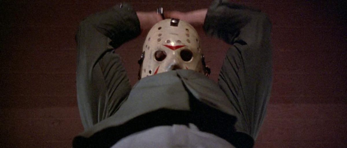 Jason Voorhees, che indossa la sua caratteristica maschera da hockey, tiene entrambe le mani sopra la testa mentre sta per affettare qualcuno in Friday the 13th Part III.